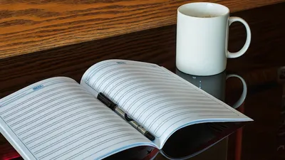 An open notebook with a pen beside a coffee mug on a reflective table.